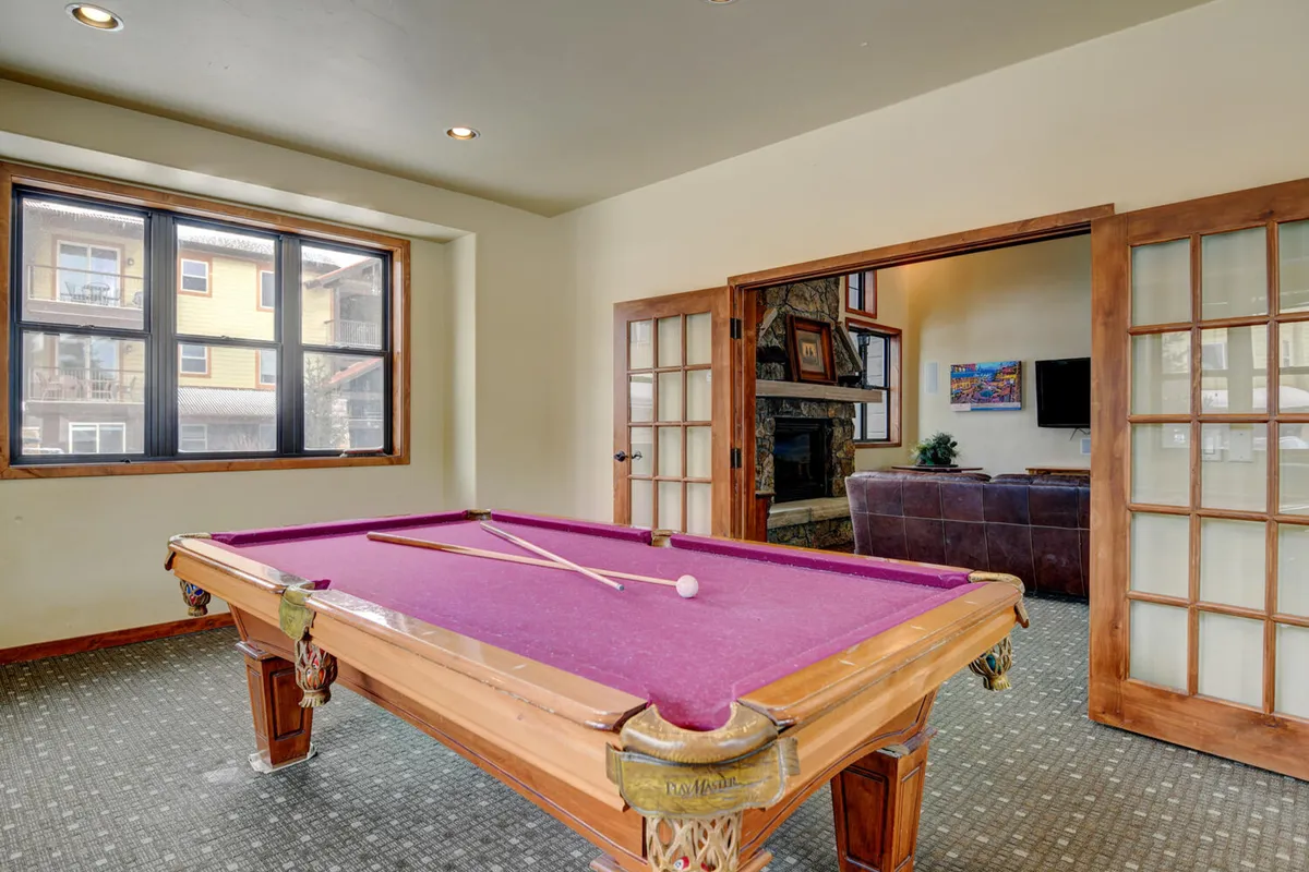 A stylish interior game room with a pool table, large windows bringing in natural light, and an open doorway to a connected living area. The space is set up for socializing and friendly competition.