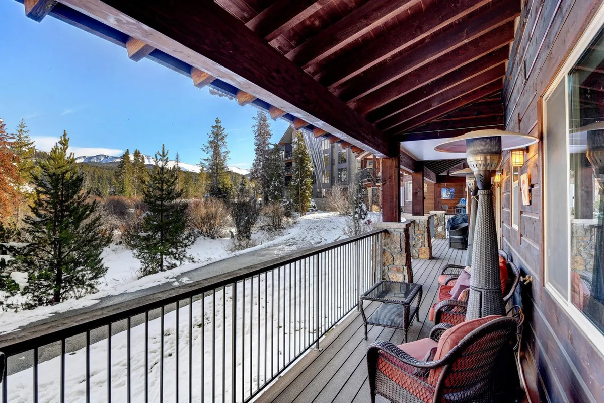 A riverside patio with wooden railing and comfortable outdoor seating overlooks a snow-covered landscape with distant mountains. The inviting scene suggests relaxation, with potential for enjoying a hot beverage while surrounded by natural beauty.