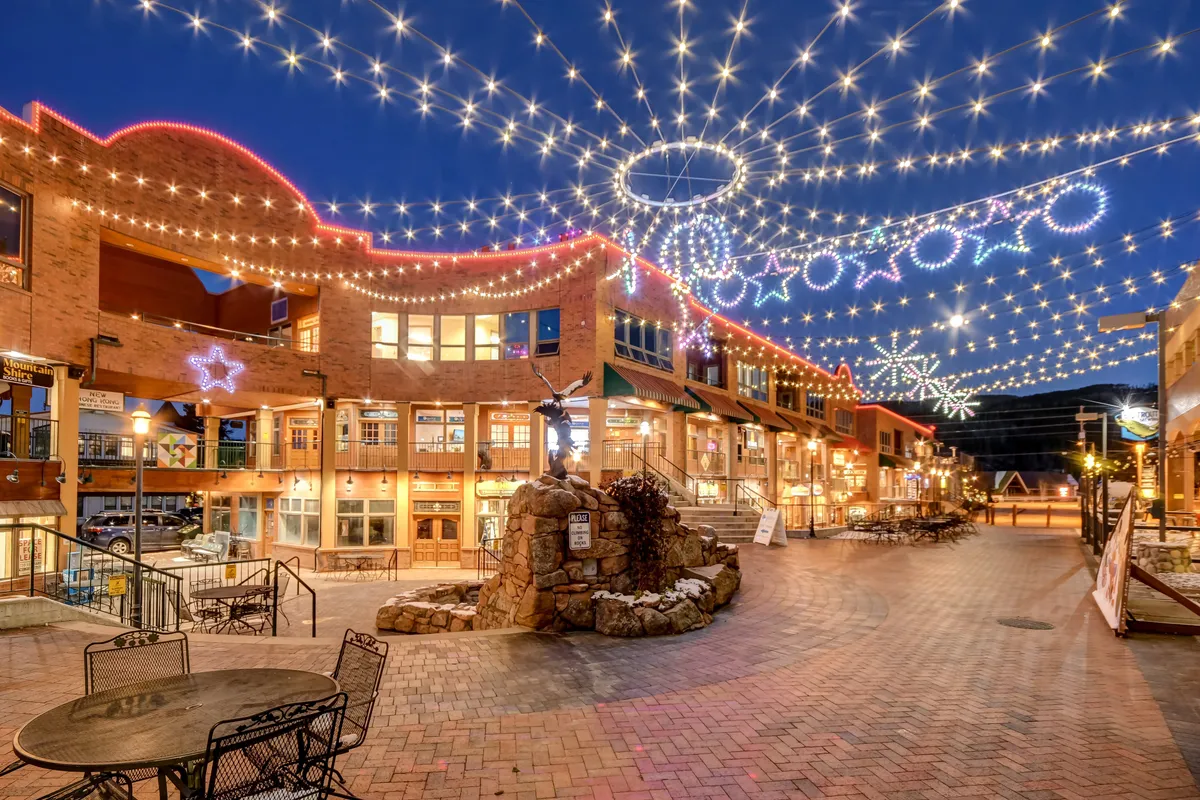 A vibrant outdoor dining area features decorative string lights, a brick building with festive touches, and a central fountain sculpture. The environment is lively, suggesting opportunities for dining out, holiday shopping, and social gatherings just steps away from the home.
