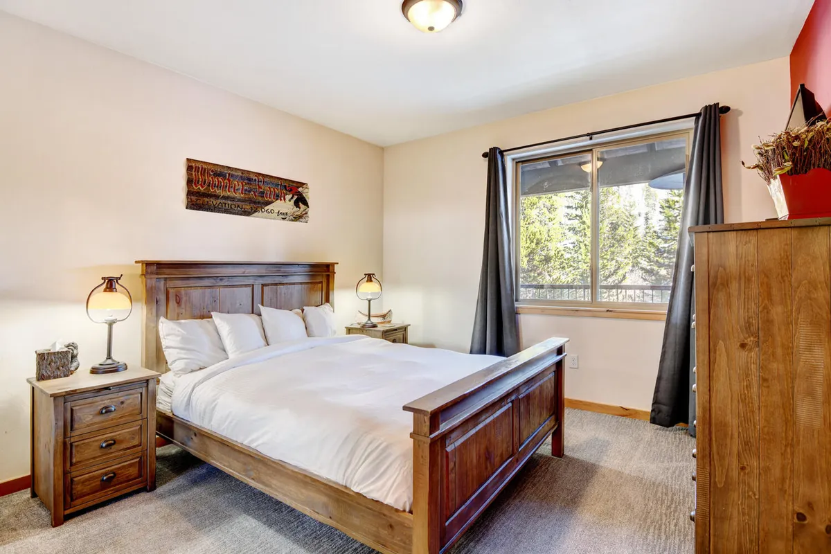 A calm and restful bedroom with a wooden bed frame, decorative wall art, matching nightstands with lamps, and a window adorned with curtains. The soft natural light and neutral tones create a peaceful environment suitable for relaxation.