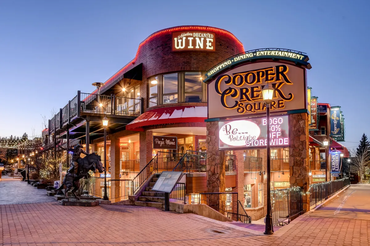 A bustling outdoor plaza featuring the Cooper Creek Square sign, inviting wine shop signage, outdoor seating areas, and a lively statue. The space is vibrant with people enjoying dining, shopping, and entertainment under clear skies.