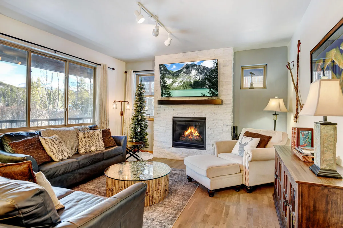 The image showcases a beautifully appointed living room with a crackling fireplace, a plush seating area centered around a stylish coffee table, and a large window framing scenic mountain views. A big-screen TV invites guests to relax and enjoy movies, while natural light fills the space, creating a warm and inviting atmosphere.