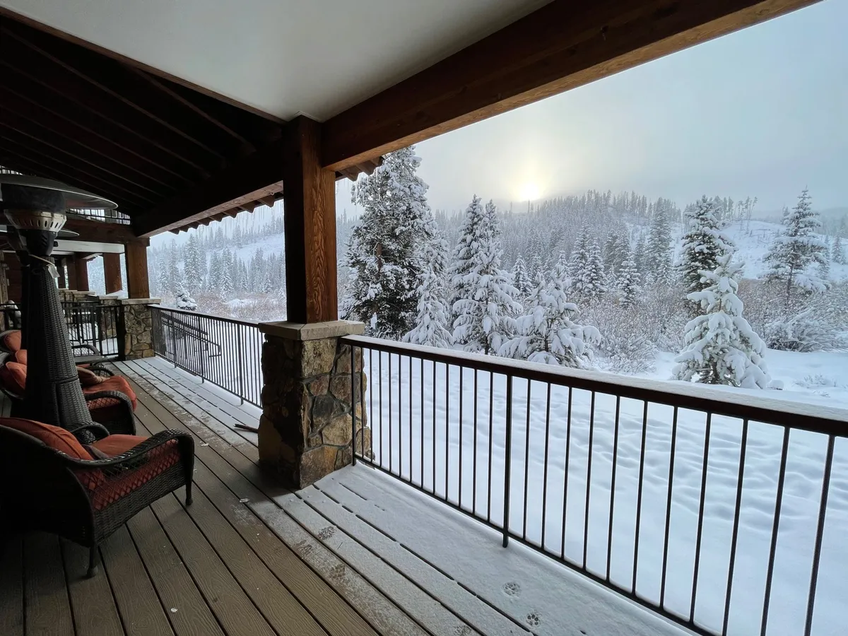 A private wooden deck with stone railings, outdoor seating, and heat lamps overlooks a snow-covered landscape and distant trees. The setting exudes warmth and comfort, inviting guests to relax outdoors even in winter. Soft sunset light filters through, creating an all-seasons ambiance.