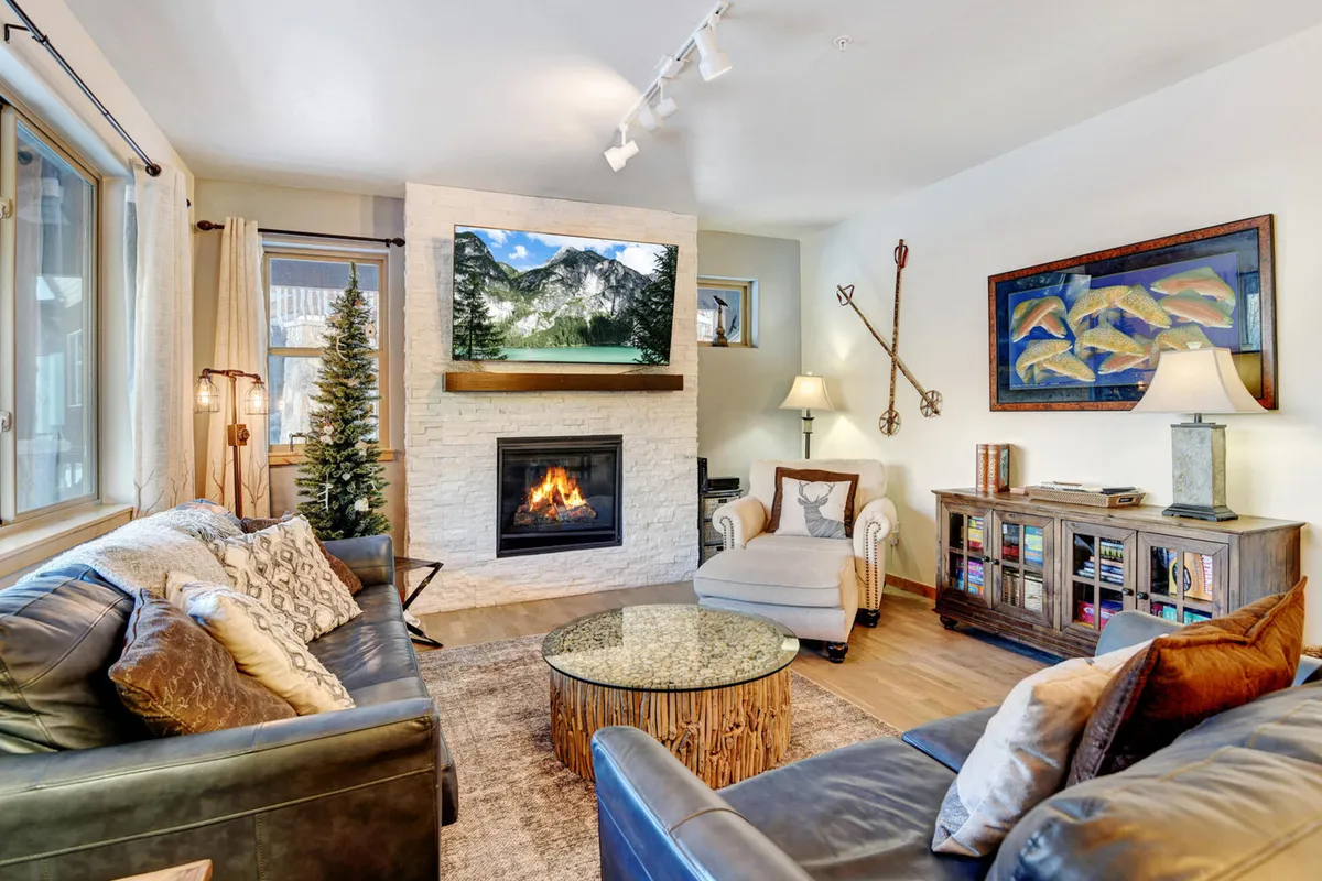 A warm, inviting living area featuring a glowing fireplace, a wall-mounted TV with mountain views, a coffee table, comfortable seating, and tasteful mountain-inspired artwork. Large windows let in abundant natural light, connecting the space to its scenic outdoor surroundings.