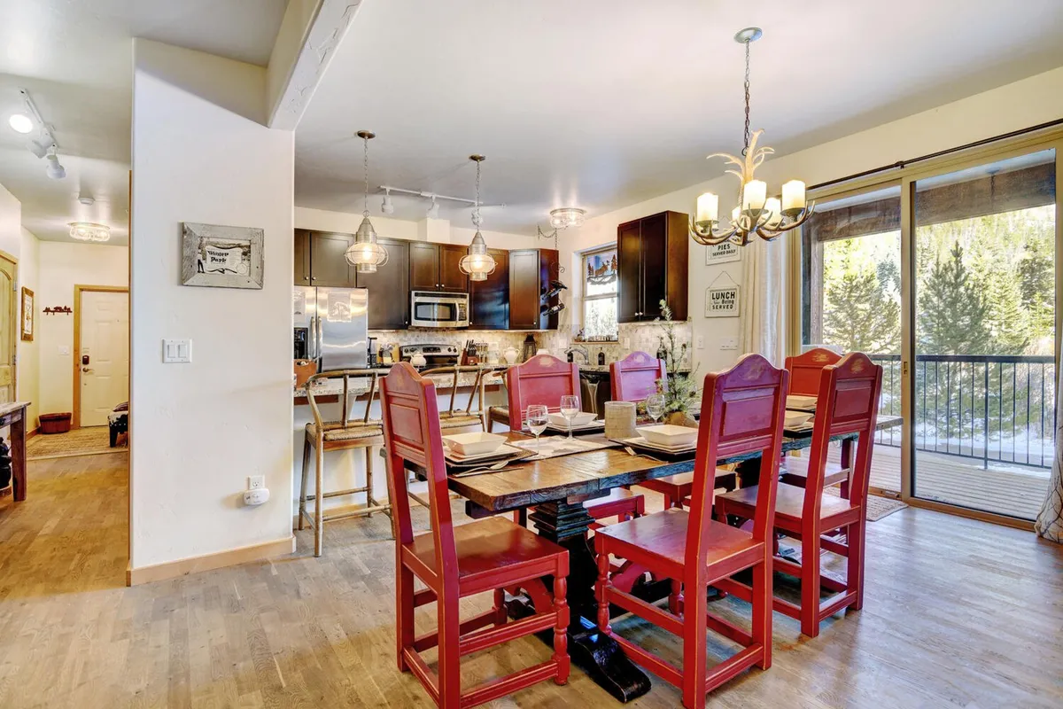 A cozy dining nook with a wooden table set for six, vibrant red chairs, an adjacent kitchen area, and large windows that fill the space with natural light. The scene suggests a warm, communal atmosphere ideal for shared meals and entertaining.