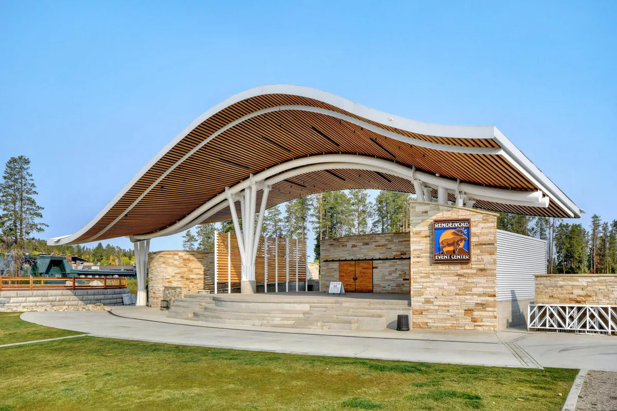 The recognizable curved wooden roof and stone facade of the local event center, with clear event center signage, set against a festive backdrop. The area is set up for concerts, community gatherings, and outdoor events.