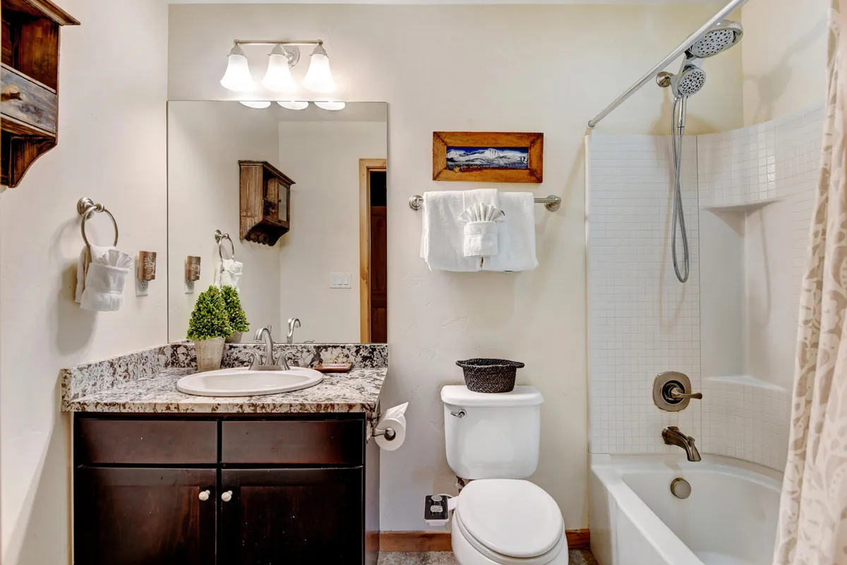 A modern bathroom featuring a granite countertop, large mirror, wooden shelf, and a well-lit shower area. The clean and refreshing space suggests high-end toiletries and a spa-like experience.