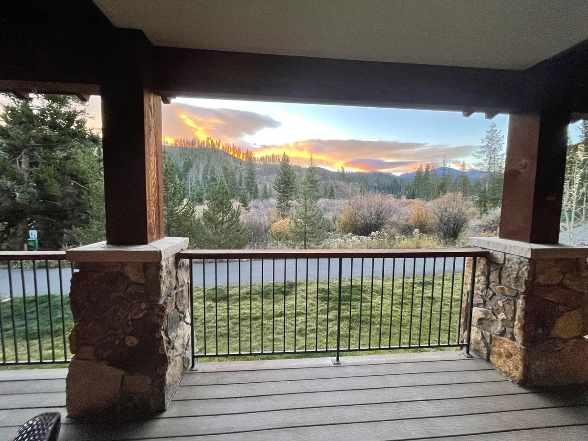 A spacious porch features stone pillars and a railing, offering panoramic views of a glowing sunset over mountain landscapes. The outdoor space is ideal for socializing, relaxing, or observing wildlife, capturing the essence of evening leisure.