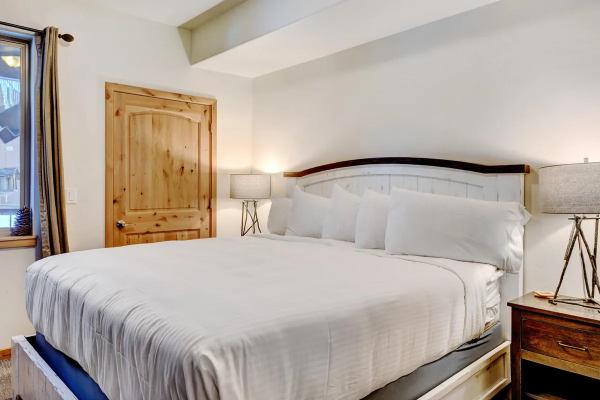 A warmly lit bedroom featuring a plush king-sized bed with premium bedding, soft lamps on either side, and a wooden door that enhances the room's rustic charm. The space radiates comfort and tranquility, ideal for restful sleep and quiet evenings.
