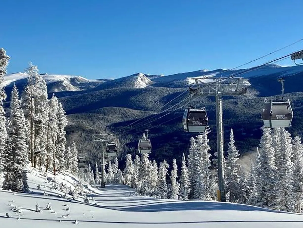 A panoramic view of a ski lift ascending into snow-covered trees with a dramatic mountain range in the background, all beneath a wide, clear sky. The untouched snow and the prominence of the ski lift evoke the excitement of winter sports and alpine exploration.